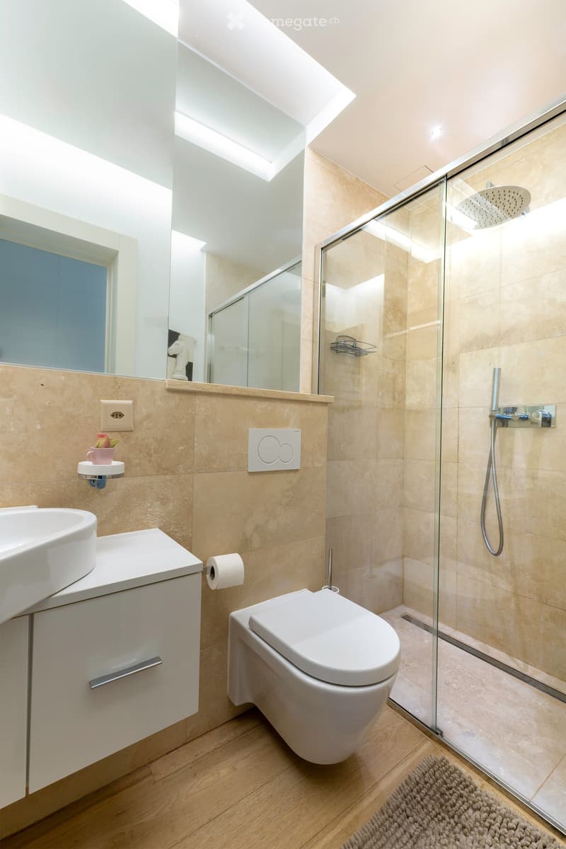Modern bathroom with a white sink, toilet, glass-enclosed shower, and a large mirror. Features beige tiles, wooden flooring, and a small decorative cup on the sink.