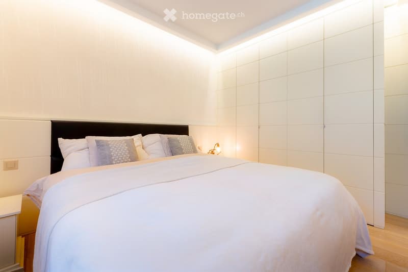 Modern bedroom with a large bed, white bedding, decorative pillows, a sleek black headboard, and a bedside lamp on a nightstand.