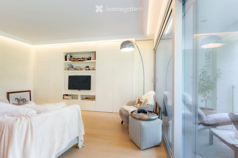 Modern living room with light wood flooring, white walls, built-in shelving, a television, a sofa, a chair, a coffee table, and a floor lamp. Large glass windows offer a view of an outdoor area with a plant.