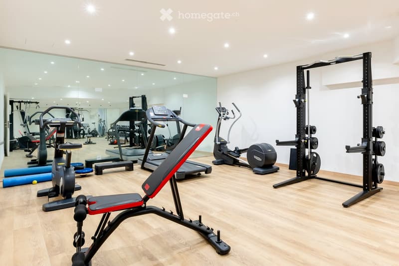Well-lit fitness room with wooden flooring, large mirror wall, and various exercise equipment including a treadmill, elliptical machine, bench press, and weight machine.