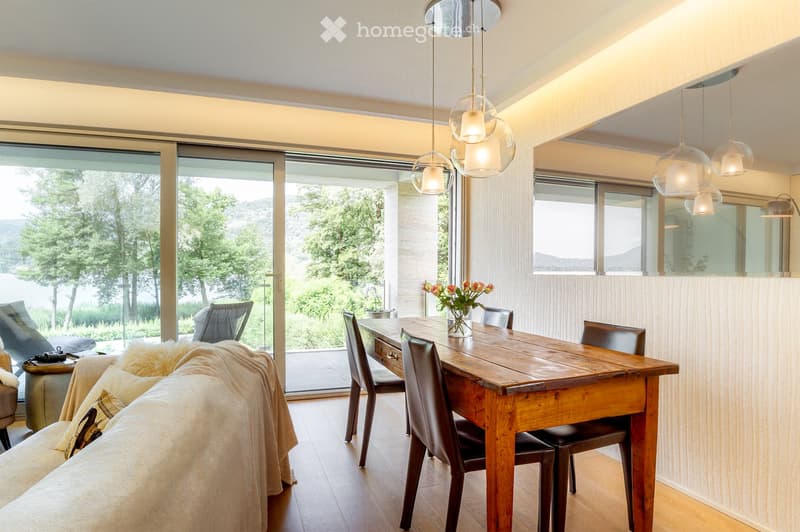 Bright, modern living and dining area with large windows offering a scenic view, a wooden dining table with chairs, a sofa with a fur throw, and a vase with flowers on the table.