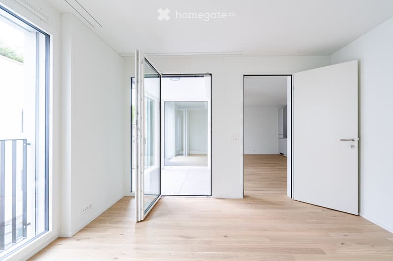 Empty hallway with light wooden flooring, white walls, and large glass doors leading to another room.