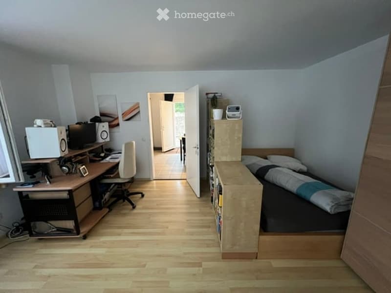 A small, well-lit bedroom with a desk, chair, bed, wardrobe, and speaker. The room features light wooden flooring and white walls.