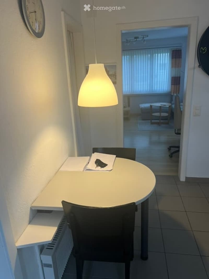 A small, modern room with a round table and chairs, a radiator, and a clock on the wall. The room is connected to another space with a bed and a desk visible in the background.