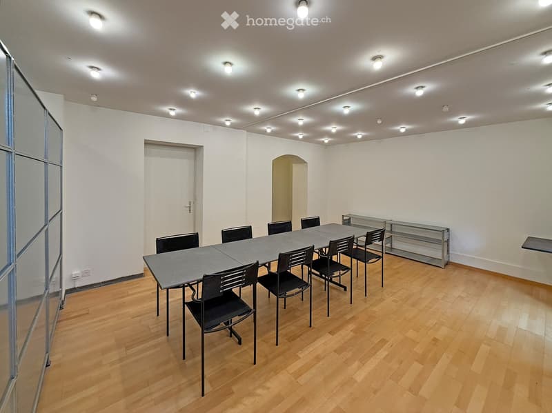 Bright, unfurnished room with wooden flooring, a long rectangular table surrounded by black chairs, and modern lighting fixtures on the ceiling.