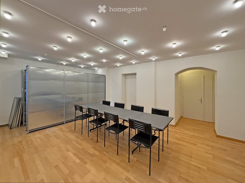 Bright, unfurnished room with wooden flooring, a long rectangular table surrounded by black chairs, and a large silver partition on one side.