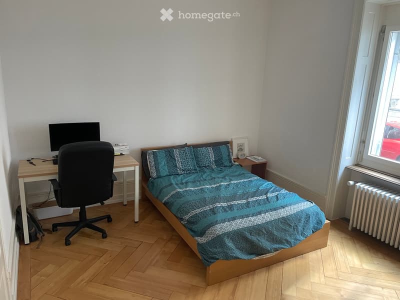 Cozy bedroom with a wooden bed, blue patterned bedding, a desk with a chair and screen, and a radiator under the window.