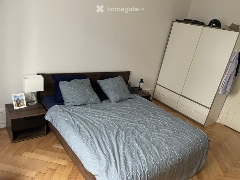 Cozy bedroom with a wooden bed, nightstand, lamp, and a large mirror wardrobe. The bed is neatly made with blue bedding and pillows.