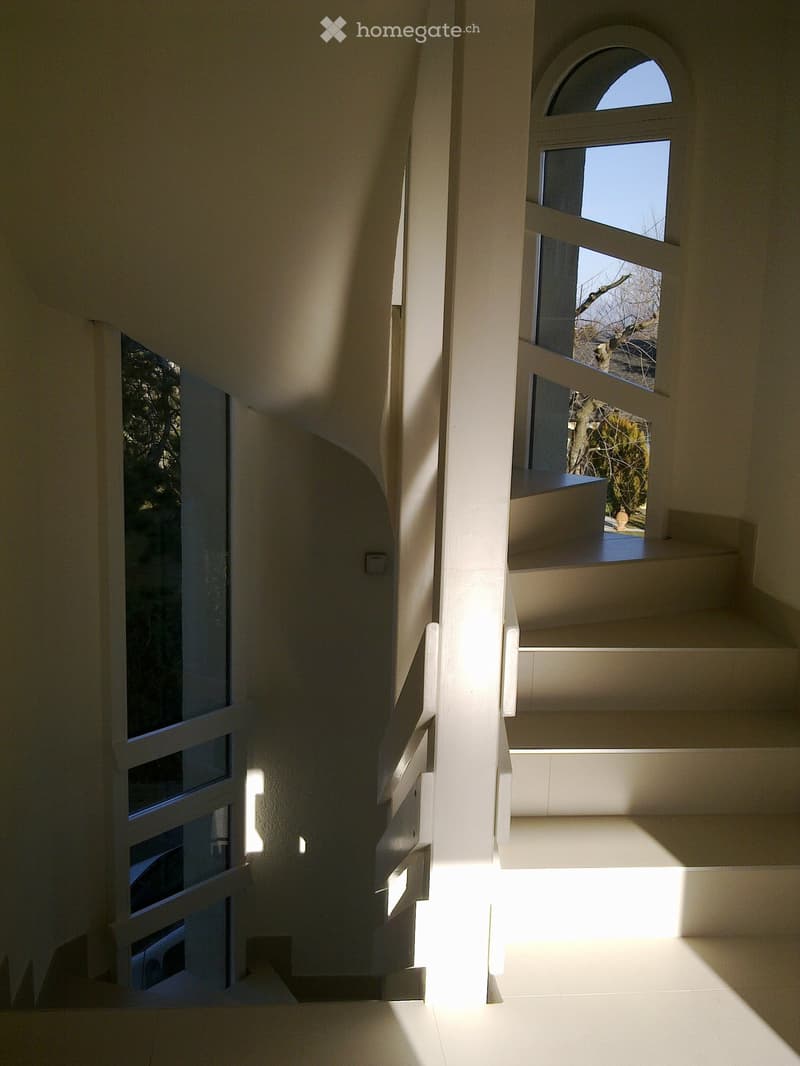 Modern, minimalist hallway with white walls, large windows, and a curved staircase leading upwards.