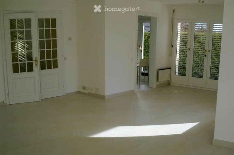 Empty room with white walls, tiled floor, and large windows with blinds. There is a small table and chair in the corner, but no other furniture.
