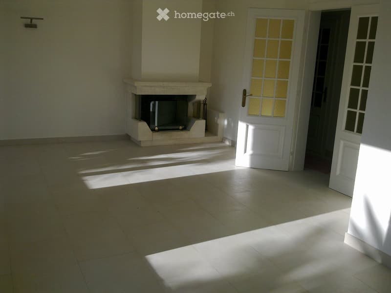 Empty room with tiled floor, white walls, and a fireplace. Sunlight streams in from an open door.