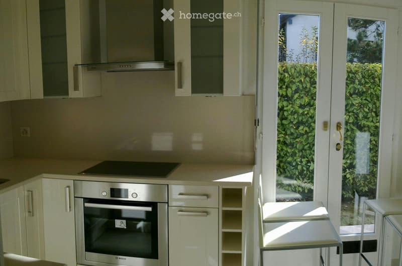 Modern kitchen with white cabinets, stainless steel oven, induction cooktop, and range hood. Features a large glass door leading to a green outdoor area.