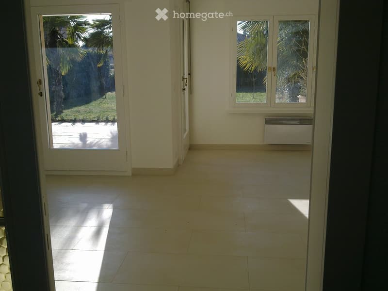 Empty hallway with tiled floor, white walls, and large windows showing a view of a garden with palm trees. An air conditioning unit is mounted on the wall.