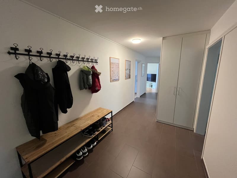 Hallway with a wooden bench, coat rack, and several jackets hanging. The hallway has tiled flooring and white walls, with a view into another room featuring a TV and a white cabinet.
