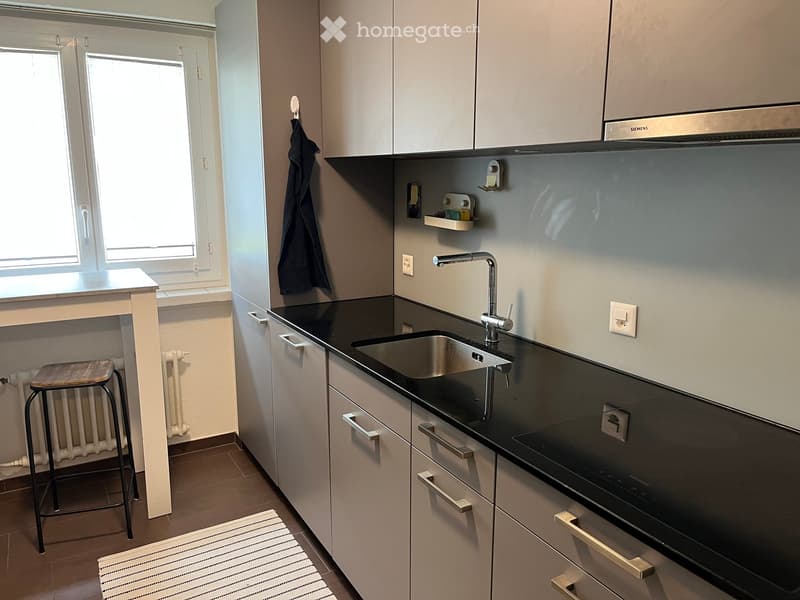 Modern kitchen with grey cabinets, black countertop, stainless steel sink, and a small wooden stool near the window.