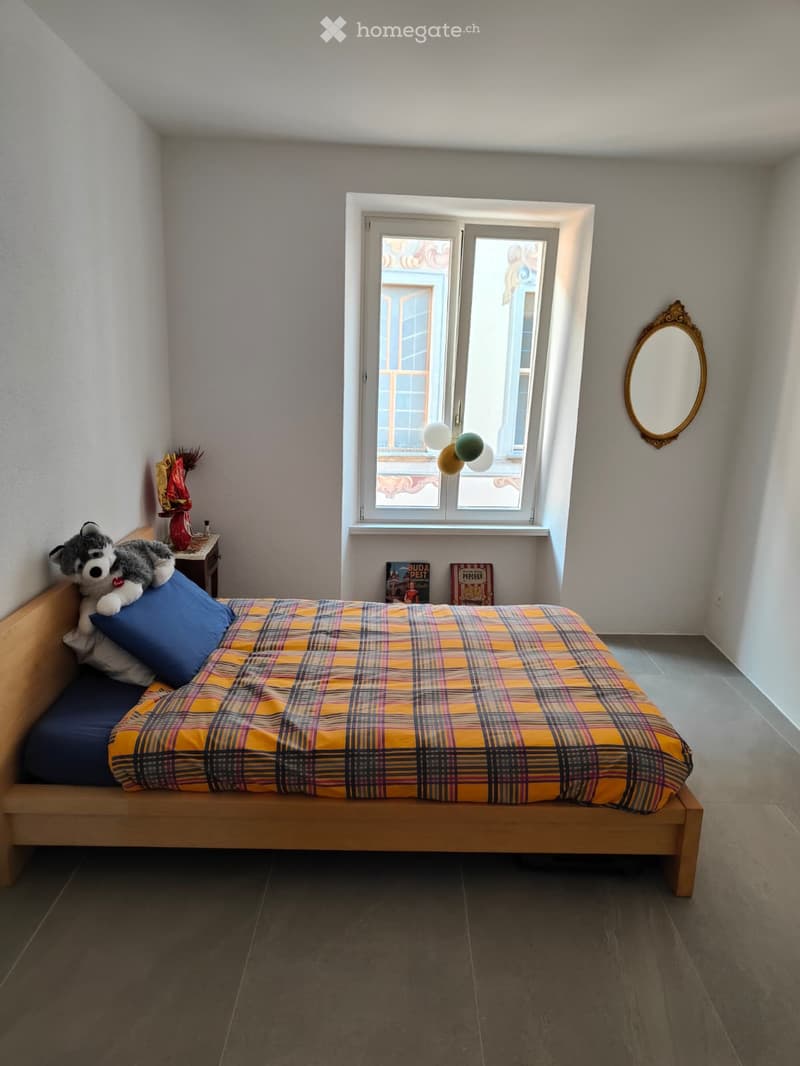 Cozy bedroom with a single bed, plaid blanket, nightstand, decorative vase, mirror, and books on the floor.