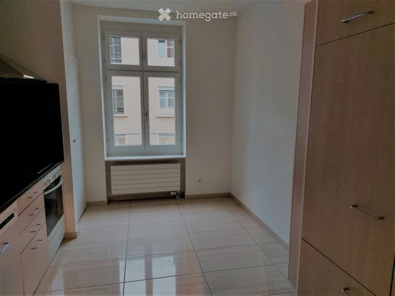 Empty room with tiled floor, light-colored walls, and a window with double doors. There is a radiator below the window and a kitchen area visible on the left side with cabinets and an oven.