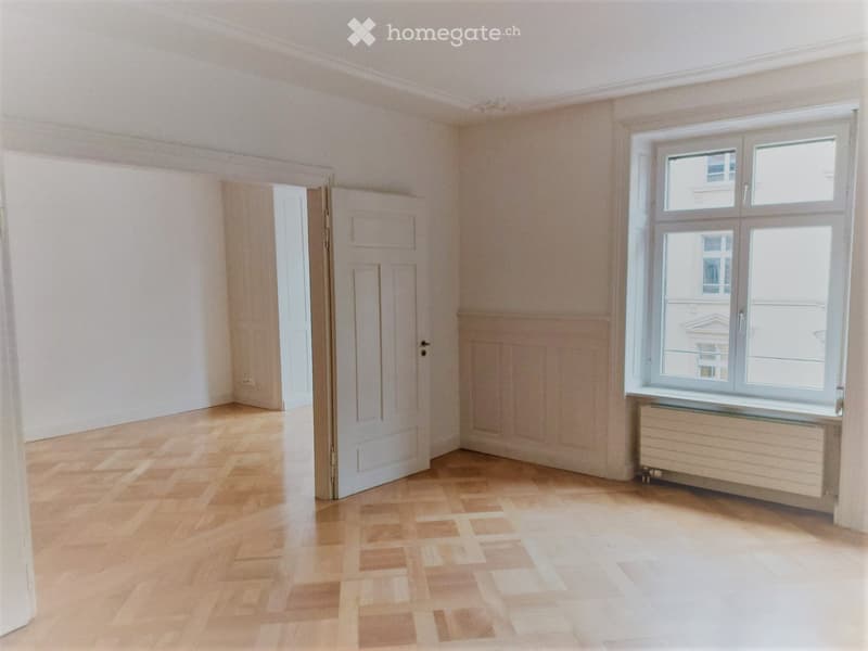 Empty room with light wooden flooring, white walls, and a large window with a radiator below it. There is a door leading to another room.