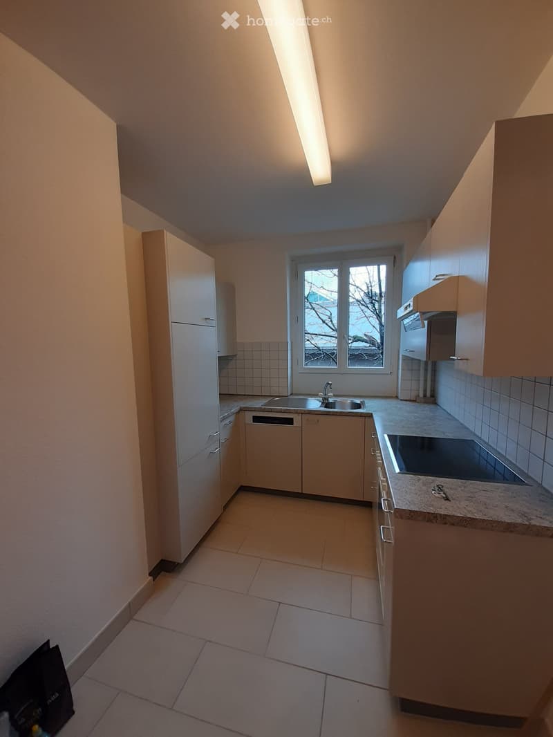 Modern kitchen with light-colored cabinets, a refrigerator, dishwasher, sink, and stove. The space features tiled walls and a window with a view of trees.