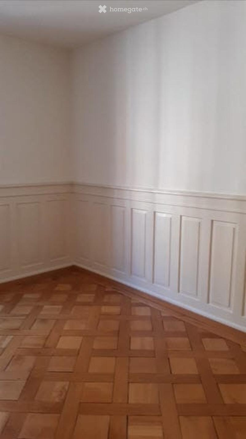 Empty room with white walls, wooden parquet flooring, and decorative paneling along the lower wall sections.