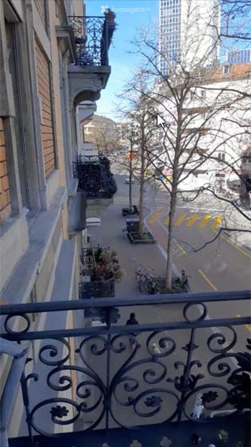 Balcony with ornate metal railing overlooking a city street, trees, and buildings in the background.