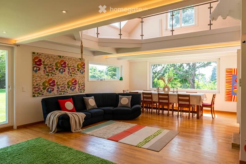 Open-concept living and dining area with wooden flooring, large windows, a black sectional sofa, a dining table with chairs, and decorative artwork.