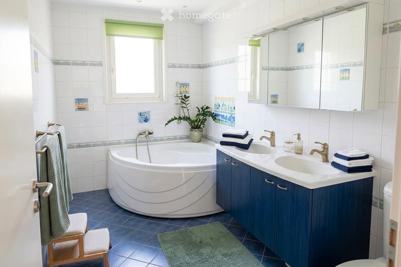 Modern bathroom with a white bathtub, double sink vanity with blue cabinets, mirror, towel rack, and decorative plant. Blue tiles on the floor and white tiles on the walls.