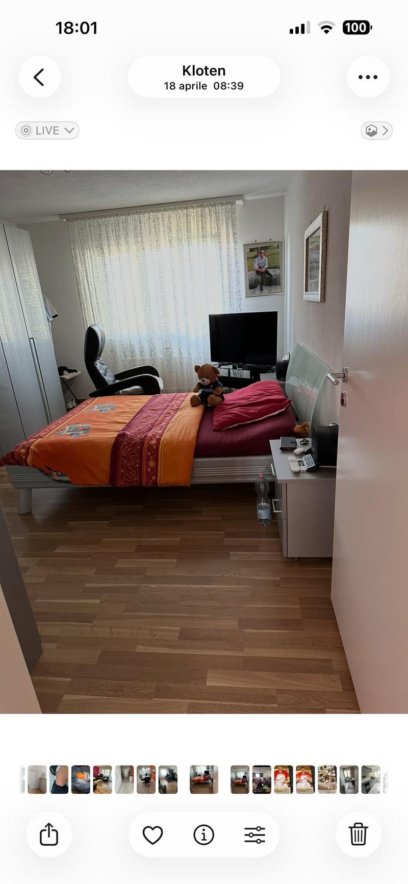A furnished bedroom with a wooden floor, a bed with colorful bedding, a chair, a small table with a screen, and a bottle on the floor.