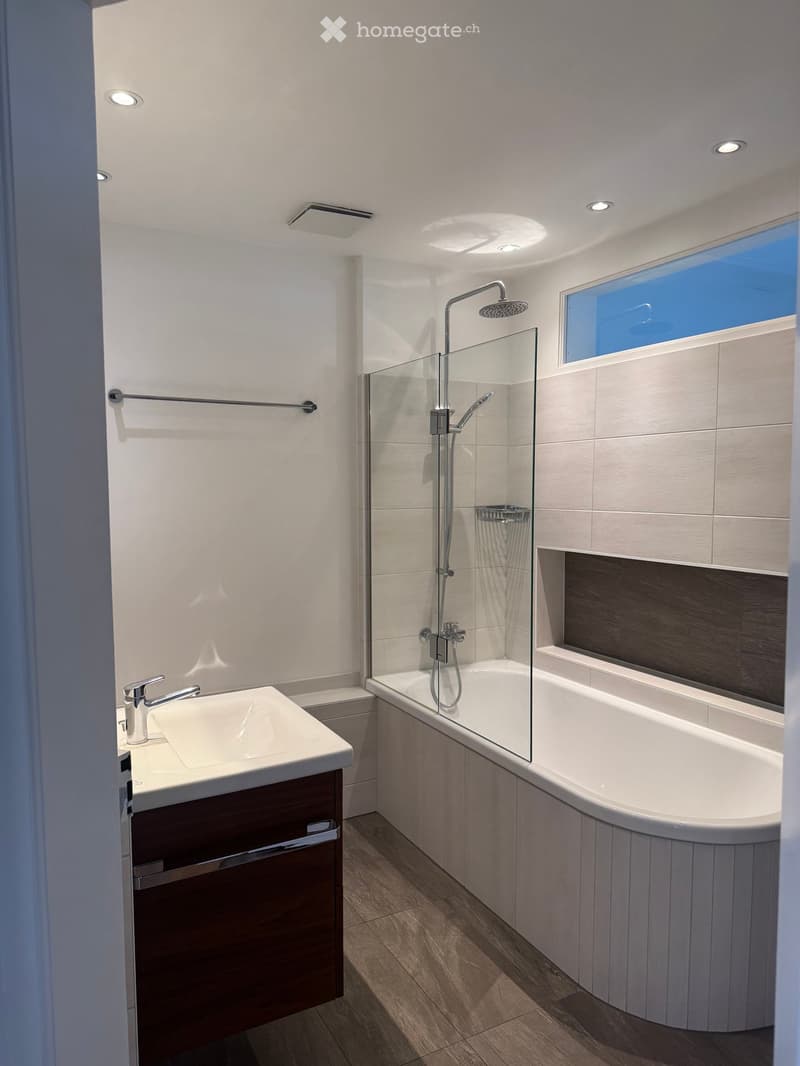 Modern bathroom with a white sink, chrome faucet, large bathtub with glass shower enclosure, and a showerhead. The room features wooden flooring and recessed lighting.