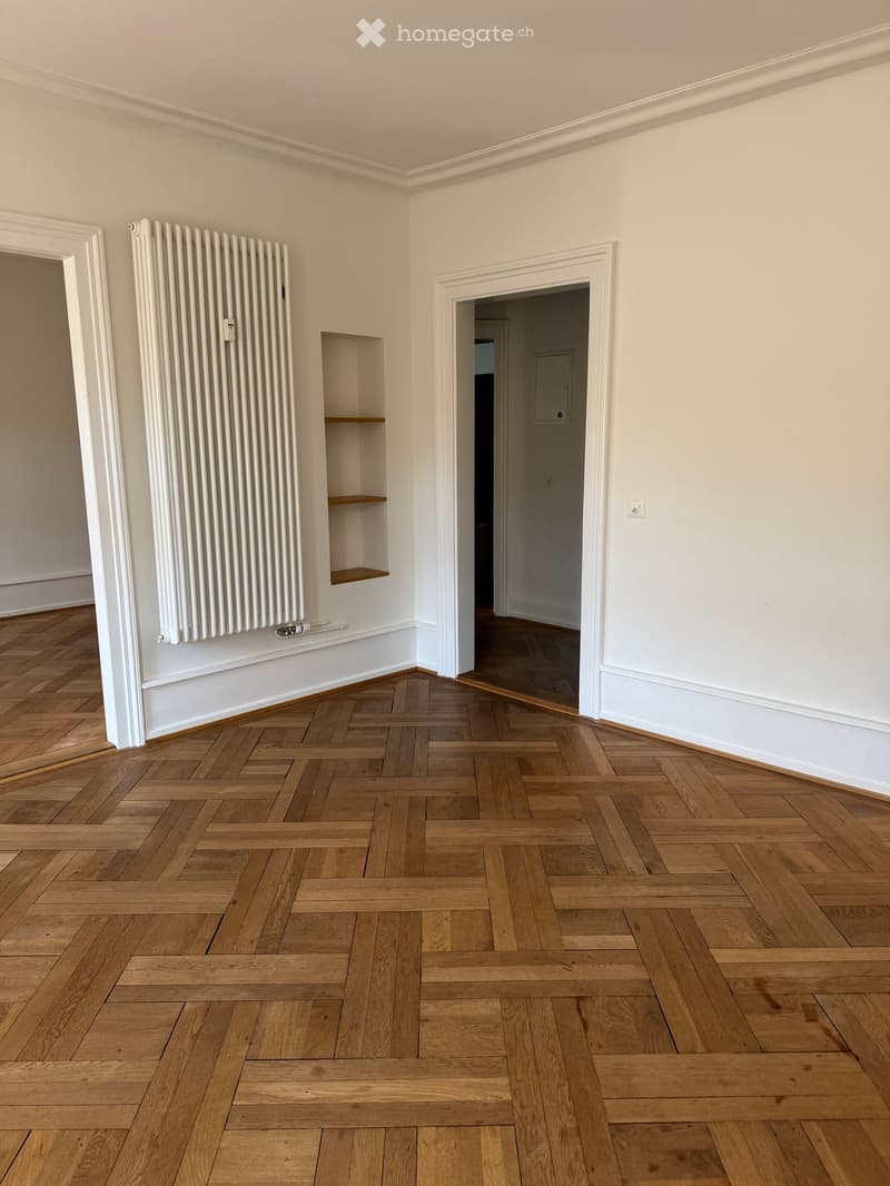 Empty room with wooden herringbone flooring, white walls, radiator, and built-in shelving unit.