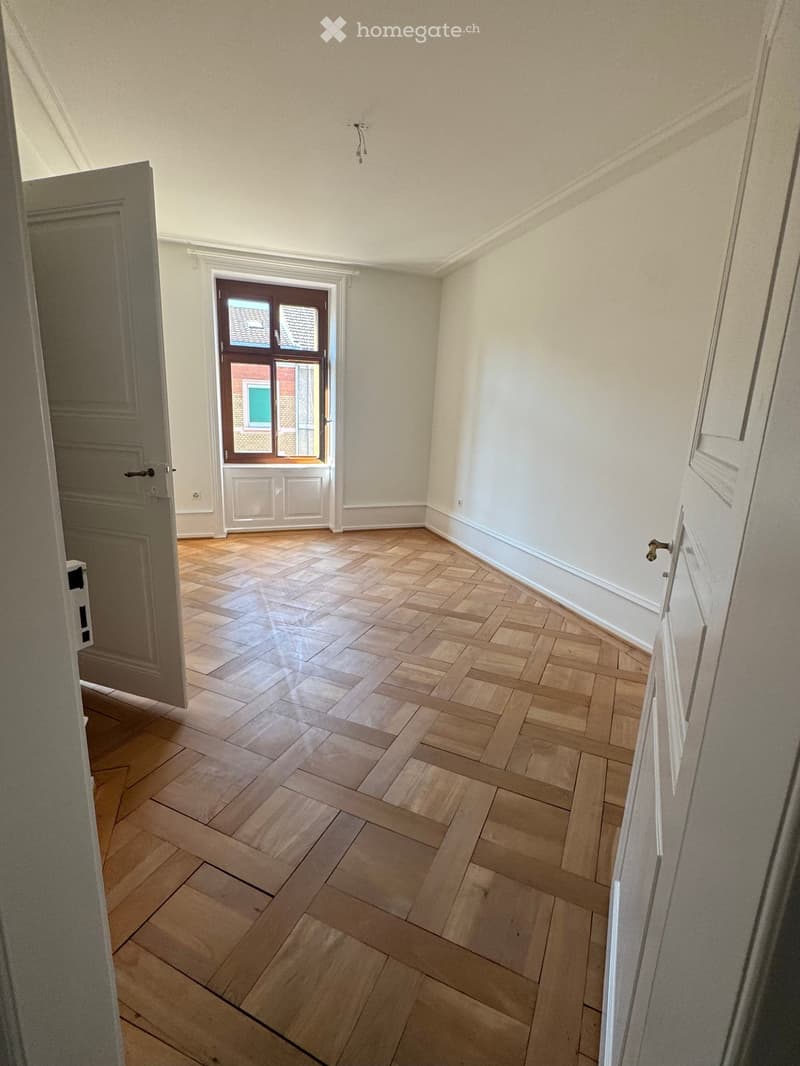 Empty room with wooden herringbone flooring, white walls, and a large window with a view of a roof and building.