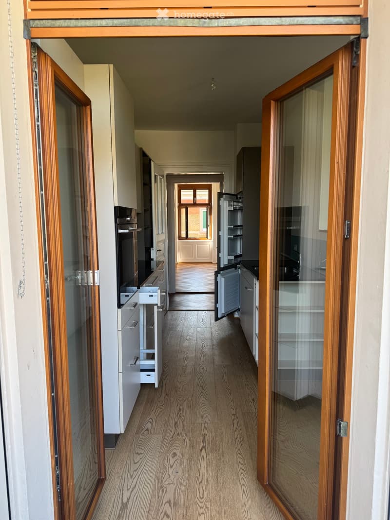 A long, narrow hallway with wooden flooring and white walls, featuring glass-paneled doors on both sides that open to adjacent rooms, including a kitchen area visible at the end.