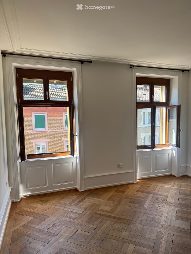 Empty room with wooden herringbone flooring, white walls, and large windows with brown frames opening to a view of neighboring buildings.