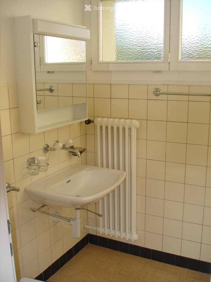A small bathroom with a white sink, faucet, radiator, and a cabinet with a mirror above it. The walls are tiled in light beige with a black baseboard.