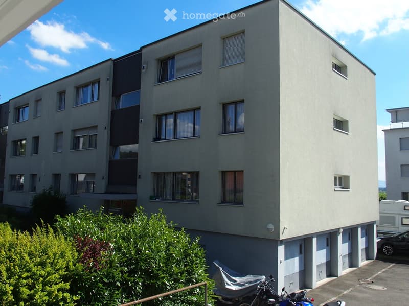 Modern multi-story apartment building with clean lines, large windows, and a covered parking area. The building features a mix of light and dark tones, with a small garden area in front.