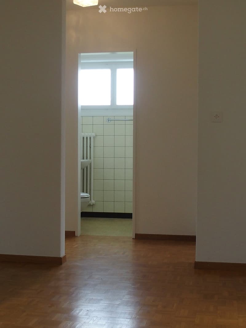 Empty hallway with wooden flooring, leading to a small bathroom with tiled walls, a toilet, and a radiator.