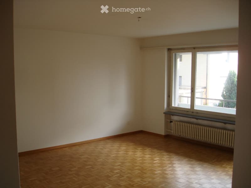 Empty room with wooden flooring, white walls, and a large window with a radiator below it.