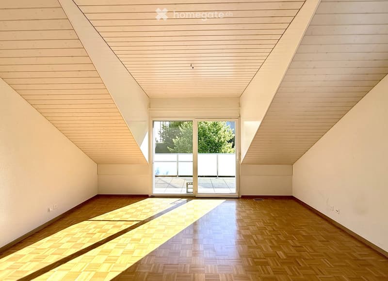 Bright, empty room with wooden flooring and slanted wooden ceiling. Large glass doors lead to an outdoor area with a tiled patio and greenery visible beyond.