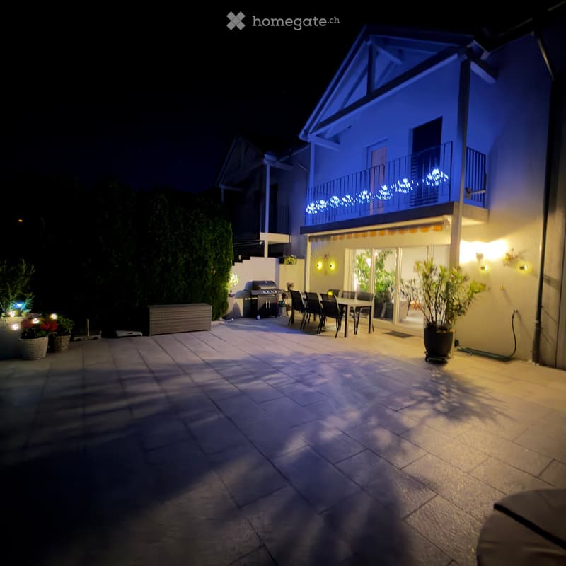 Well-lit outdoor patio with a dining set, grill, potted plants, and string lights on the balcony of a modern house.