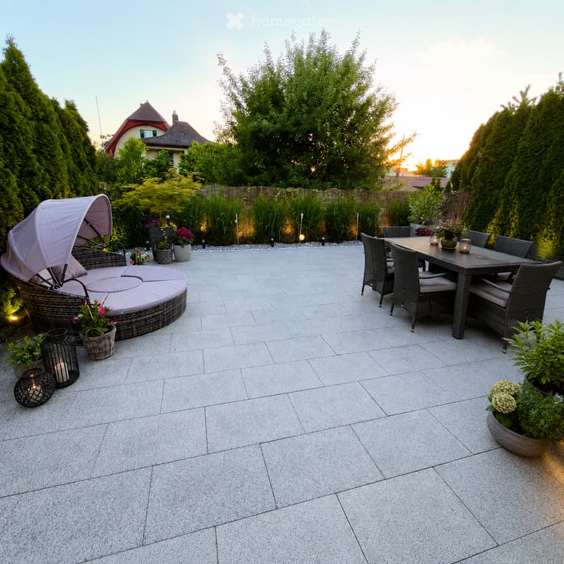 Well-maintained patio with grey stone tiles, featuring a round wicker sofa with cushions, a rectangular dining table with four chairs, and several potted plants. The area is bordered by a wooden fence and trees, with ambient lighting and a view of a house