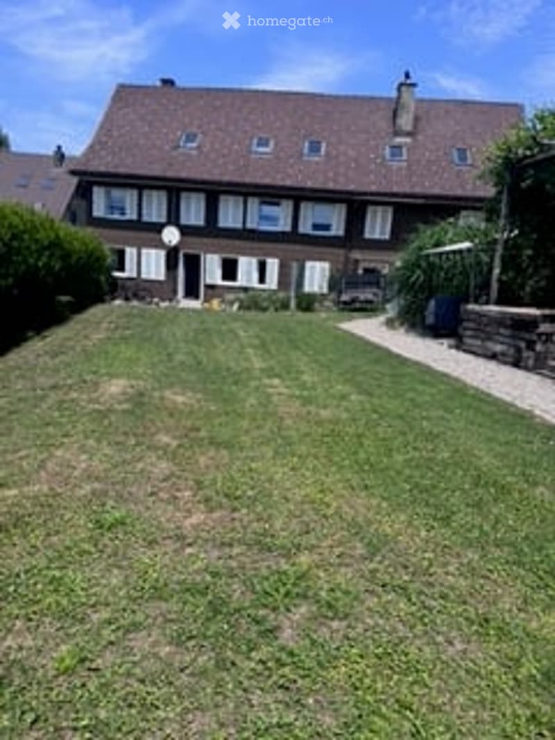 Well-maintained front lawn with trimmed hedges, a satellite dish on the roof, and a small patio area with a table and chairs near the house entrance.