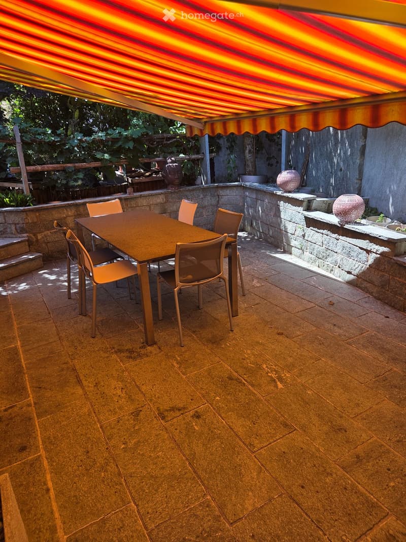 Outdoor patio with stone flooring, a table and chairs, and a covered awning. There are decorative items and plants in the background.