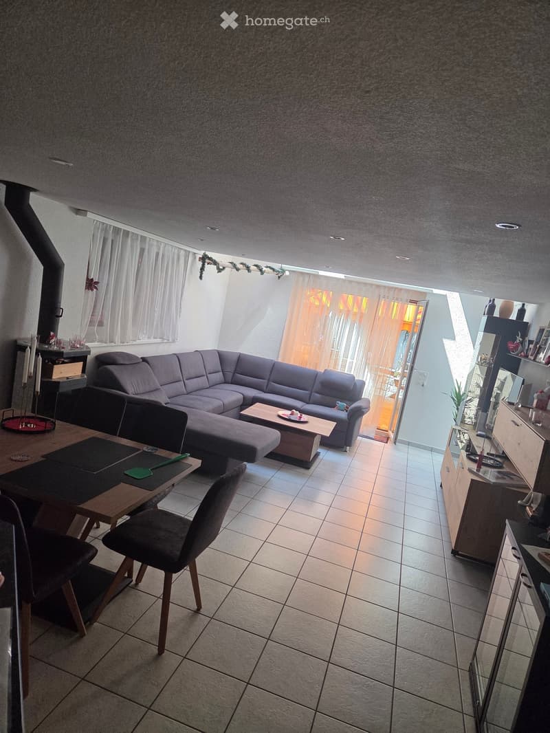 Living and dining area with a grey sectional sofa, coffee table, dining table with chairs, and a small cabinet with a mirror. The space features tiled flooring and large windows with curtains.