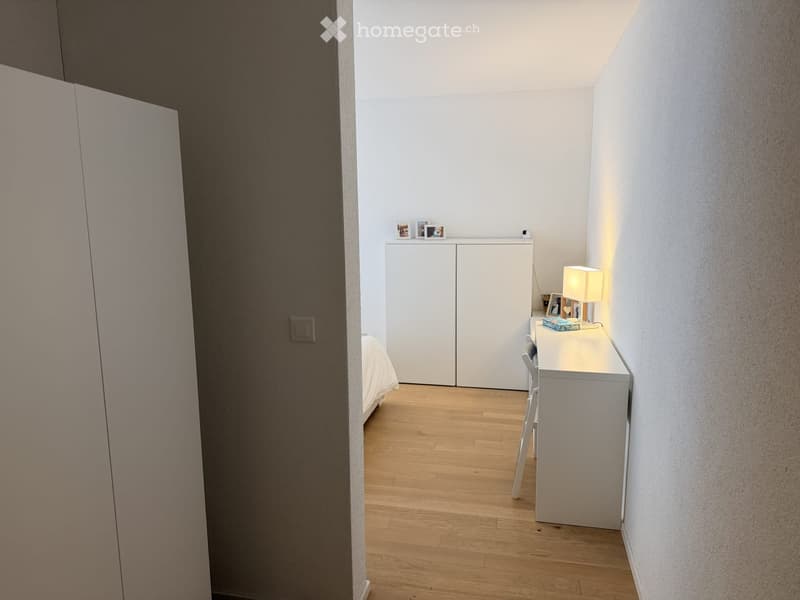 Modern, minimalist bedroom with light wooden flooring, white walls, built-in wardrobe, small desk with chair, and a lamp on the desk.