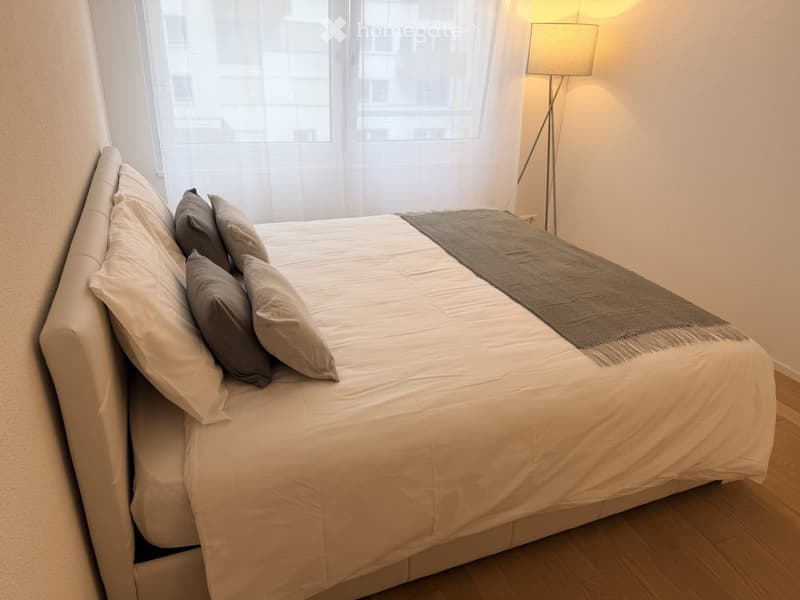 Modern bedroom with a large bed, multiple pillows, a grey blanket, and a floor lamp near a window with sheer curtains.