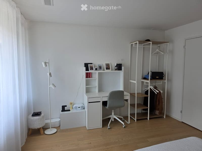 A modern, well-lit room with a white desk, ergonomic chair, floor lamp, and shelves holding books and decorative items. The room features wooden flooring and a large window with white curtains.