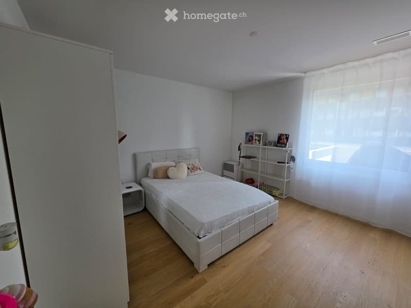 Modern bedroom with a white bed, nightstand, and a shelf with picture frames and books. Light wooden flooring and white walls.