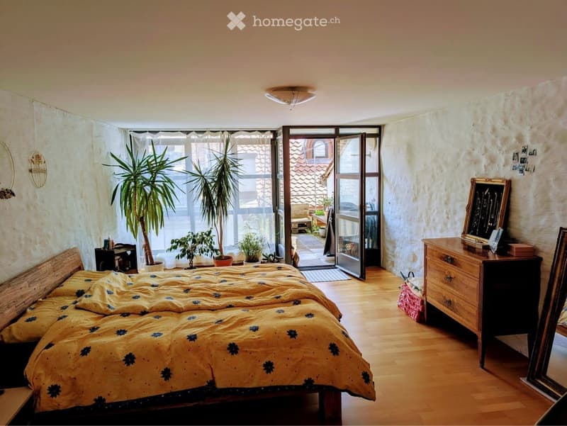 Cozy bedroom with wooden flooring, large bed with yellow and blue bedding, nightstand, dresser, potted plants, and a sliding glass door leading to a balcony.