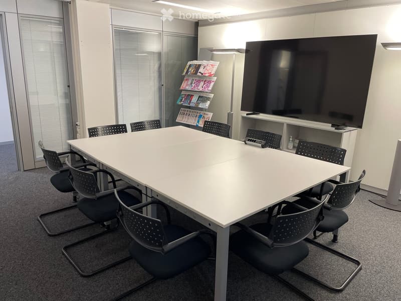 Furnished Room. Conference room with a large white table, black chairs, a large screen, and a magazine rack.