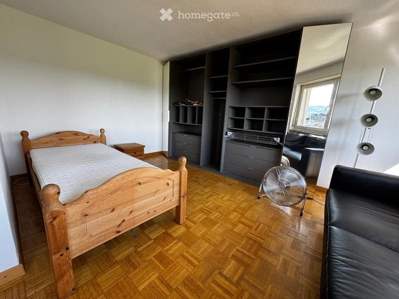 A well-lit bedroom with a wooden bed, a grey sofa, a fan, a large mirror, a nightstand, and a built-in bookshelf with drawers.